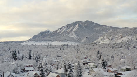 Low-drone-shot-moving-backward-of-Boulder-Colorado-and-rocky-Flatiron-mountains-after-large-winter-snow-storm-covers-trees,-homes,-streets,-and-neighborhood-in-fresh-white-snow