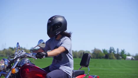 Man-rides-a-red-motorcycle-down-a-farm-road-in-the-summer-wearing-a-helmet-with-a-field-in-the-background
