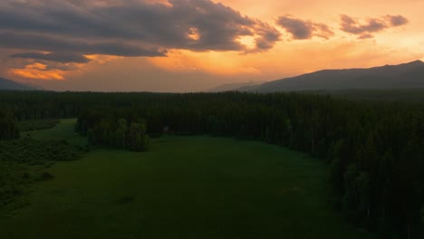 Aerial-drone-flying-through-open-lush-green-field-surrounded-by-tall-trees-in-Montana-mountains-during-stunning-orange-and-yellow-cloudy-sunset