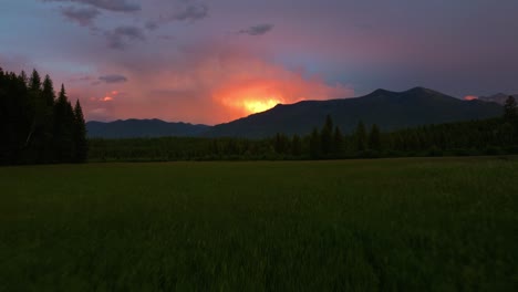 Epic-summer-sunset-over-Montana-mountain-landscape-during-rain-storm-with-lightning-bolt-flashing-through-the-clouds-as-camera-pulls-back-over-lush-green-field-of-grass-lined-with-tall-trees