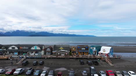 Drone-flyover-of-quaint-fishing-town-in-Homer-Alaska,-fisherman's-wharf-on-coast-line-with-mountain-range-in-background