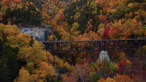 Young-person-in-a-yellow-jacket-explores-a-train-track-bridge-on-the-side-of-the-mountain-during-the-autumn-with-fall-foliage-in-the-background