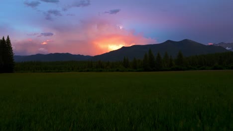 Epic-summer-sunset-over-Montana-mountain-landscape-during-rain-storm-with-lightning-bolt-flashing-through-the-clouds-as-camera-pushes-in-over-lush-green-field-of-grass-lined-with-tall-trees
