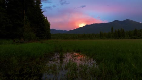 Atemberaubender-Sonnenuntergang-über-Der-Berglandschaft-Von-Montana-Während-Eines-Regensturms-Mit-Einem-Blitz,-Der-Durch-Die-Wolken-Blitzt,-Während-Die-Kamera-über-Ein-Sumpfiges-Grünes-Grasfeld-Zurückkehrt,-Das-Von-Hohen-Bäumen-Gesäumt-Ist