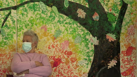 Elderly-woman-sitting-with-a-protection-mask-on-her-face,-the-back-wall-painted-with-a-tree-alluding-to-autumn-colors