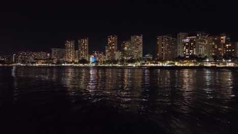 30fps-nighttime-drone-footage-of-waikiki-hawaii-flying-into-strip-with-skyline-and-traffic-in-honolulu-with-waves-and-city-lights-and-palm-trees