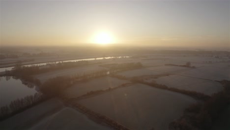 Drone-flying-backwards-with-the-camera-tilting-down-slowly-filming-winter-farmfields-at-sunrise-in-4k