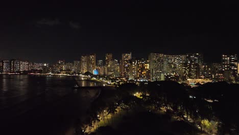 reveal-drone-aerial-night-shot-of-waikiki-showing-strip-and-city-lights-downtown-in-evening-behind-trees