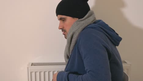 Freezing-young-man-with-hoodie,-hat-and-scarf-sitting-next-to-a-heater-and-shivering-with-cold,-energy-crisis