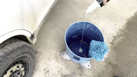 Pouring-a-little-bit-of-car-soap-shampoo-into-a-bright-blue-bucket-with-a-microfiber-washing-mitt-on-top-and-a-filthy-white-car-with-all-terrain-tires-next-to-it