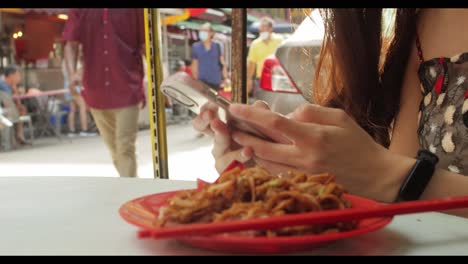 Asiatisches-Hübsches-Mädchen,-Das-Am-Telefon-SMS-Mit-Essen-Auf-Dem-Tisch-Schreibt,-Während-Die-Leute-Tagsüber-In-Malaysia-Chinatown-Vorbeigehen
