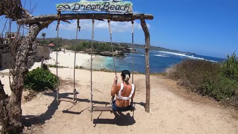 Girl-on-a-swing-with-a-beer-in-her-hand-on-a-beach-in-Nusa-Lembogan,-Bali,-Indonesia