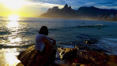 Woman-sitting-on-the-rocks-at-Arpoador-Beach-at-sunset-with-the-iconic-mountains-of-Rio-de-Janeiro-in-silhouette