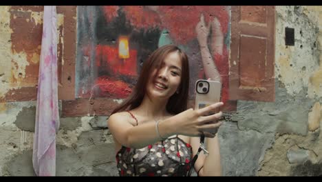 Una-Chica-Guapa-Asiática-Sonríe-Y-Saluda-En-Una-Videollamada-En-Una-Pared-Mural-En-El-Barrio-Chino-De-Malasia-Durante-El-Día