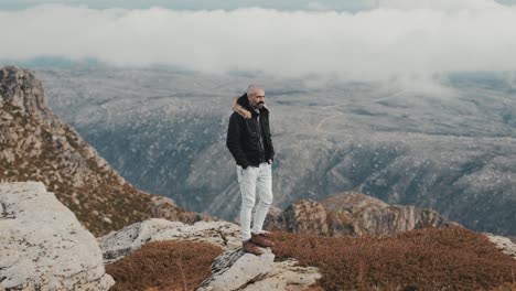 A-handsome-man-with-short-hair-and-a-mustache-stands-on-top-of-a-rock,-hands-in-pockets,-and-surveys-the-stunning,-mountainous-landscape-with-foggy-clouds-on-the-horizon