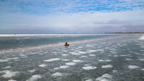 Man-fishing-on-a-frozen-lake,-Lake-St-Clair,-Mitchell's-Bay,-60fps-aerial-drone-shot,-with-icy-lake-texture-and-windmill-background