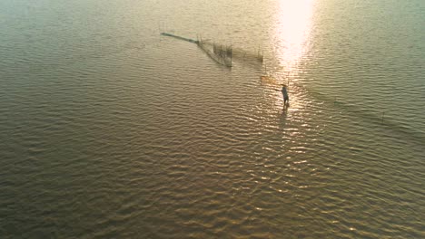 Fisherman-cast-net-fishing-on-the-vast-expansive-Tonle-Sap-waterway,-Asia,-Aerial-birds-eye-view-of-silhouetted-angler-throwing-his-net-during-late-afternoon-setting-golden-sun,-slow-mo,-copy-space