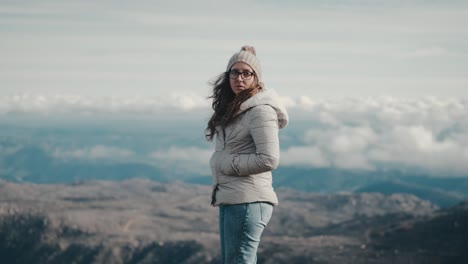 Young-thoughtful-woman-in-a-gray-jacket,-hat,-and-scarf-contemplates-a-stunning-mountain-landscape-on-a-hike