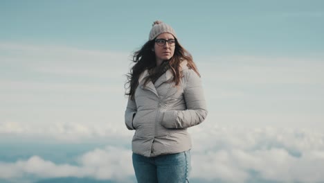A-beautiful-young-woman-in-a-gray-jacket-and-glasses-contemplates-the-beauty-of-nature-while-taking-in-the-stunning-panoramic-views-of-the-mountains-and-clouds