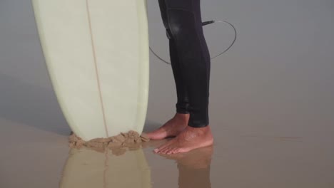 Low-angle-of-Surfer-in-wetsuit,-barefoot-on-wet-sandy-beach-floor-with-white-surfboard,-Handheld-shot