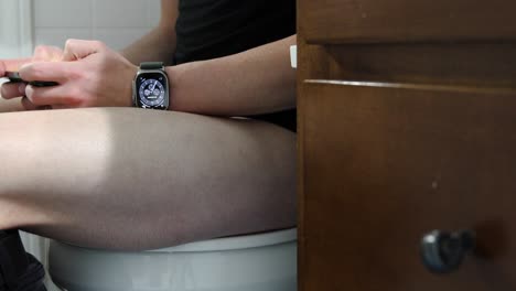 Junger-Weißer-Mann,-Der-Auf-Einer-Toilette-Sitzt-Und-Süchtig-Nach-Seinem-Handy-Und-Seiner-Smartwatch-Ist