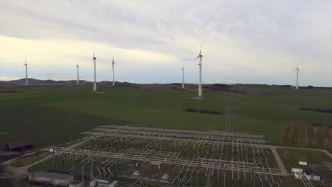 Aerial-Backwards-Shot-of-Critical-Energy-Production:-Rotating-Windmills-on-a-Field-with-a-Power-Substation-in-the-Foreground-in-Sauerland,-Germany