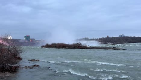 Bewölkte-Niagarafälle-Kanada-Entlang-Des-Flusses-Mit-Blick-Auf-Die-Skyline-Im-Winter