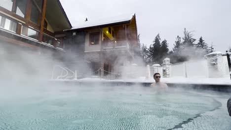 Wind-blowing-steam-off-of-a-hot-pool-swirling-at-Halcyon-Hot-Spring-Resort-in-British-Columbia,-Canada-with-a-young-white-male-with-glasses-relaxing-in-the-steam-and-the-Resort-in-full-view