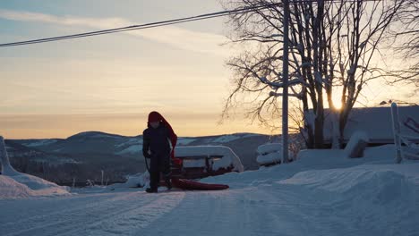 Young-Boy-Wearing-Long-Red-Hat-Getting-Off-Sled-And-Running-Back-With-Calm-Orange-Sunset-Sky-In-Background-In-Norway
