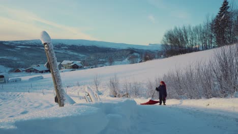 Shot-of-a-little-boy-walking-back-along-the-snow-covred-slope-enjoying-sledding-using-red-plastic-shovel-sled-in-Beitostolen,-village-in-Innlandet-county,Valdres,-Beito,-Norway-at-daytime
