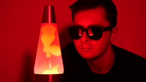 Close-up-of-white-yeen-male-looking-at-a-red-lava-lamp-while-wearing-shades
