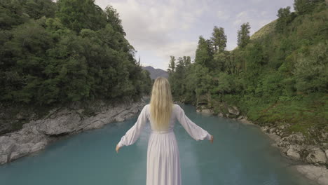 Blond-model-in-white-dress-raising-arms-slowly-while-looking-at-vibrant-blue-river