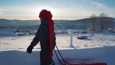 Kleiner-Junge-Mit-Langem-Roten-Hut,-Der-Schlitten-über-Die-Winterliche-Schneelandschaft-In-Norwegen-Zieht,-Mit-Strahlender-Sonne-Am-Horizont