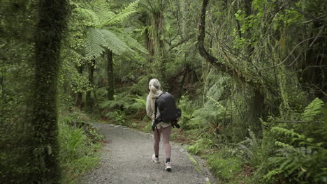 Blond-female-photographer-with-backpack-hiking-on-gravel-trail-through-lush-native-bush,-New-Zealand