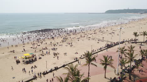 Drone-shot-of-large-crowds-on-Durban-beachfront-in-South-Africa,-people-in-the-Indian-Ocean-swimming-in-the-waves