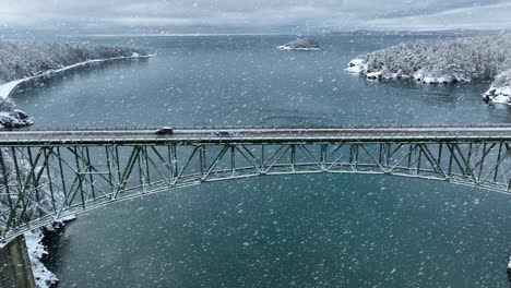 Drone-shot-of-a-car-driving-across-a-bridge-in-the-winter-with-snow-actively-falling