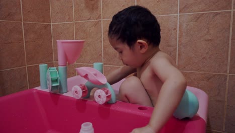 A-three-year-old-Asian-Chinese-boy-bathing-inside-a-small-plastic-pool,-filling-up-and-pouring-water-from-containers,-splashing-water,-observing-the-water-he-is-playing-with