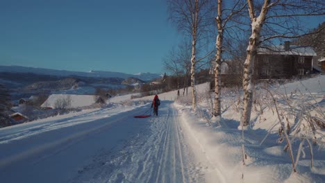 Young-Boy-Wearing-Long-Red-Hat-Running-Across-Winter-Snow-Covered-Path-Pulling-Sled-Behind-Him-In-Norway