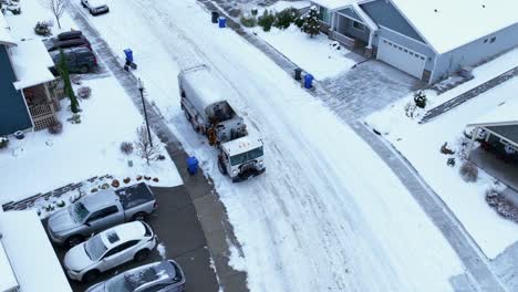 Drone-shot-of-a-garbage-truck-picking-up-trash-in-the-ice-cold-elements-with-snow-covering-the-ground