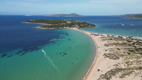 Windsurfer-surfing-along-sandy-beach-towards-peninsula-island-at-Porto-Pollo,-Sardinia---4k-Aerial-Pedestal-Up