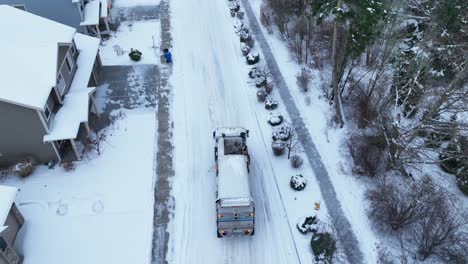 Drone-shot-following-a-dump-truck-as-it-drives-through-snow-filled-streets-to-pick-up-trash