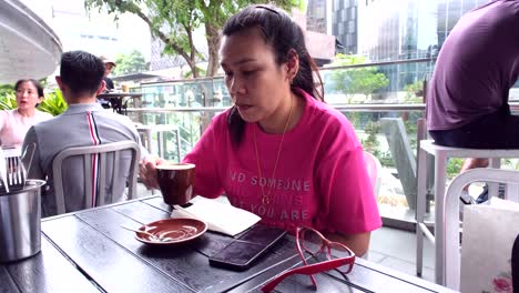 Eine-Asiatische-Frau,-Die-Tasse-Kaffee-Schlürft,-Während-Sie-Am-Tisch-Sitzt,-Und-Ein-Telefon-An-Der-Seite