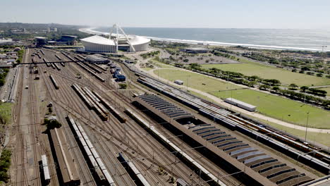 Aerial-shot-of-Passenger-train-leaving-Durban-train-station-in-South-Africa-with-Moses-Mabhida-Soccer-stadium-and-Indian-Ocean-in-background