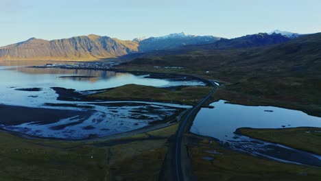 Atemberaubende-Luftaufnahme-Der-Wunderschönen-Isländischen-Straße,-Die-Sich-Entlang-Des-Sees-Mit-Majestätischem-Berghintergrund-Schlängelt,-Island