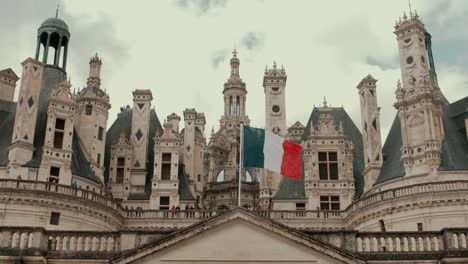 Low-angle-static-view-of-Chambord-Castle-facade-with-French-flag-waving-in-cloudy-day