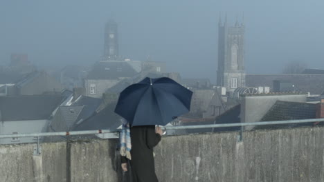 Woman-walks-with-blue-umbrella-looking-from-rooftop-towards-city-church-towers
