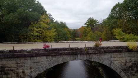 Aerial-of-a-young-man-walks-on-a-gravel-path-on-a-bridge-over-a-river-in-the-fall