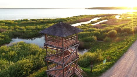 Empty-wooden-lookout-tower-near-lake-and-gravel-road-during-golden-sunset,-aerial-orbit-view