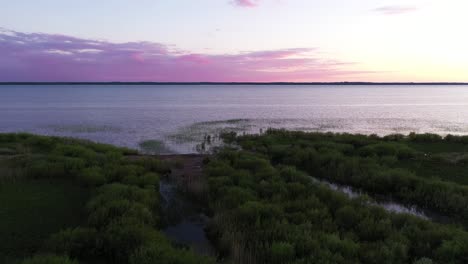 wetland-marsh-coastline-aerial-view-flying-across-calm-still-ocean-under-purple-sunset-skyline