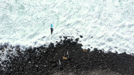 4k-Drone-top-view-shot-of-a-surfer-jumping-in-the-water-with-his-surfboard-from-the-rocky-coastline-in-Lennox-Head,-Australia